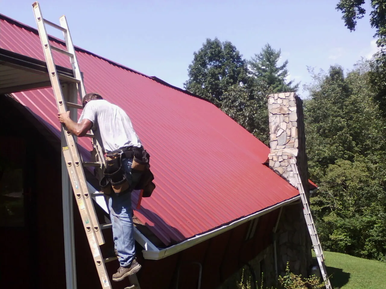 Roofer on a ladder beside a red metal roof and stone chimney — Soffit & Fascia Repair by Roofing Brothers in Spurger